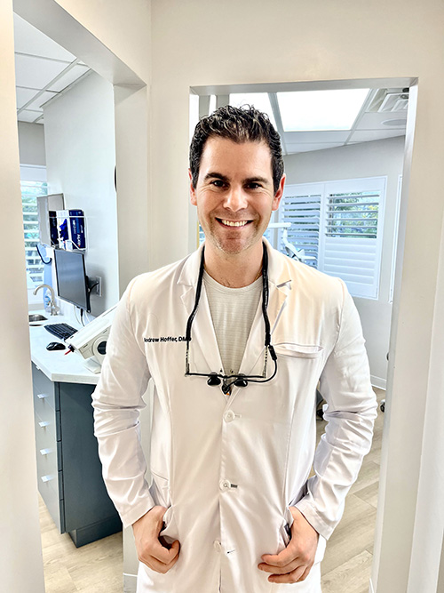 The image is a photograph of a man posing for the camera, smiling slightly and looking directly at it. He has short dark hair, facial stubble, and is wearing a light-colored shirt with a collar. The background appears to be an indoor setting with neutral colors.