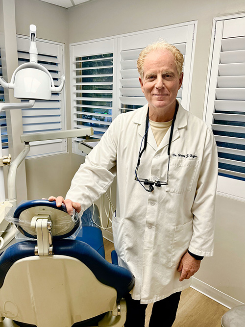 The image shows a man in a white lab coat standing indoors, smiling and looking directly at the camera.