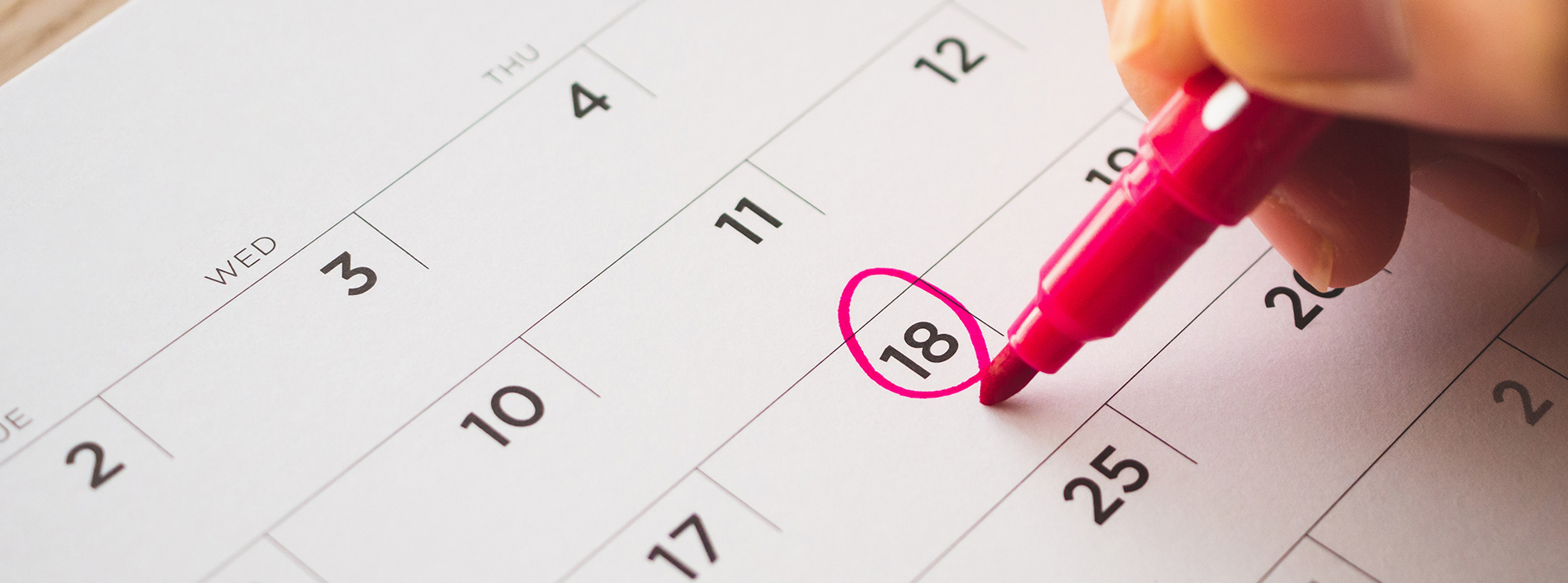 A close-up of a hand writing on a pink highlighter marker, highlighting the number 18 in a calendar with a blurred background.