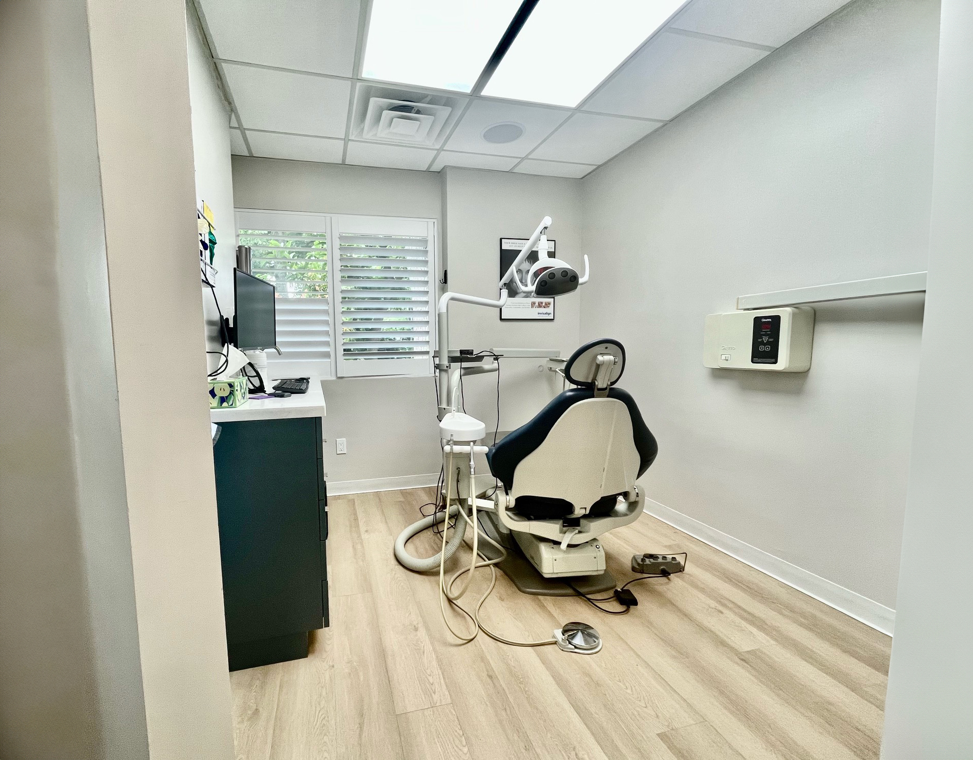 The image shows an interior space, likely a dental or medical office, featuring a dental chair and equipment in the foreground with a window to the left allowing natural light into the room.