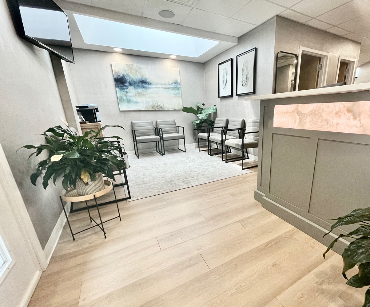 The image shows an interior view of a well-lit office space, featuring a reception area with a desk and chairs, a potted plant, a framed picture on the wall, and a whiteboard.