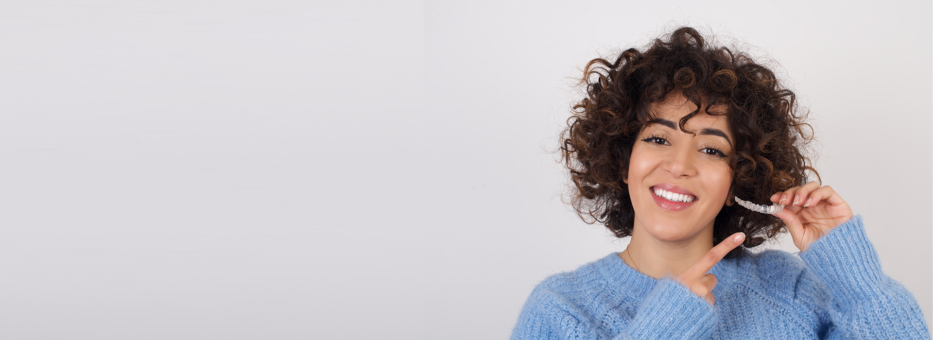 A woman with curly hair is smiling and holding a phone to her ear. She is wearing a blue top and has short, dark hair.