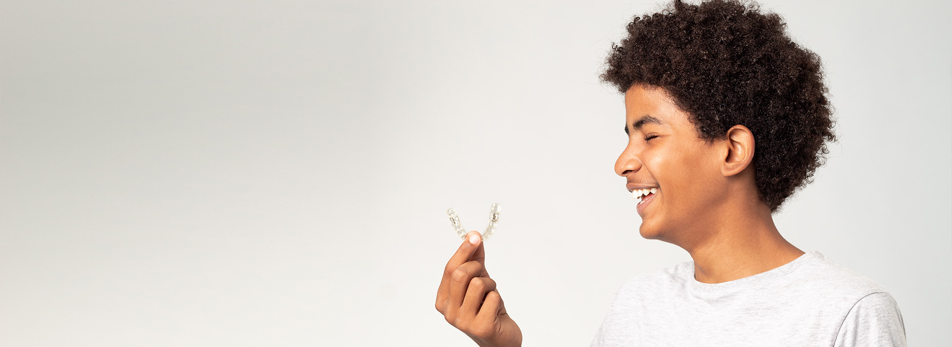 The image is a photograph of a person smiling and holding out their hand with an object that appears to be a small piece of paper or fabric. The individual has short hair, and the background is plain and light-colored, which provides contrast to the subject.