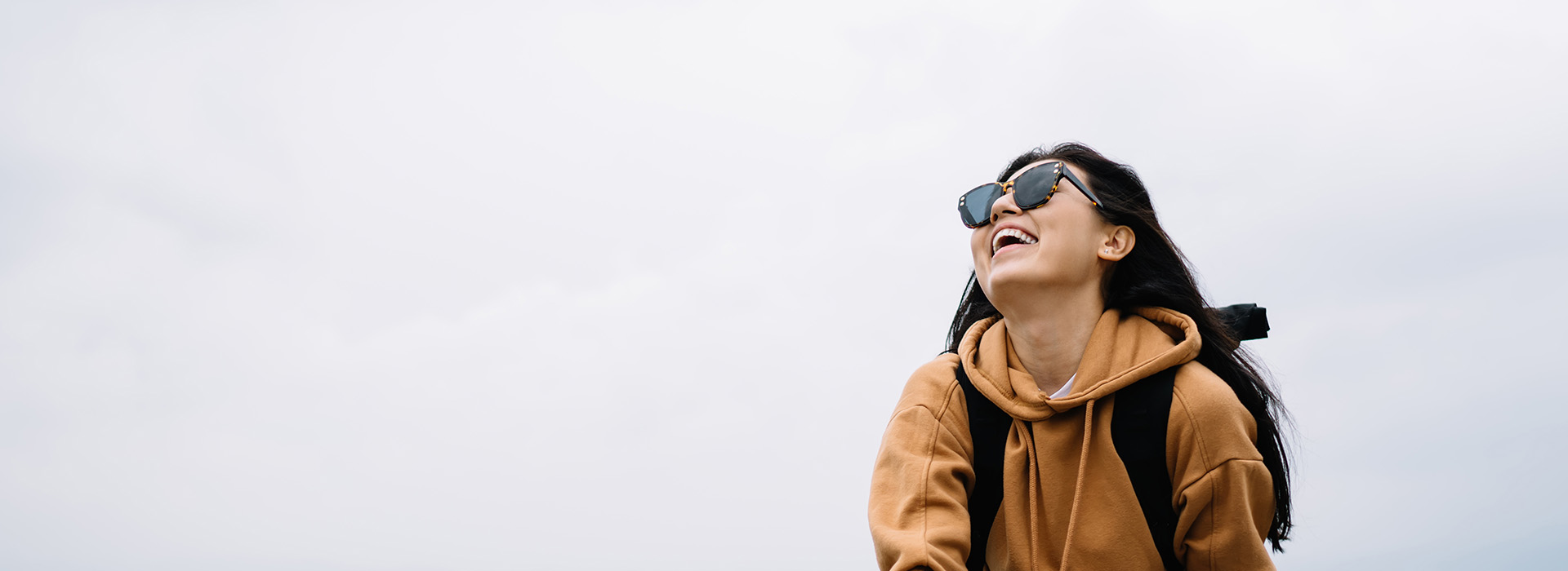 A person wearing sunglasses and a backpack is standing outdoors with a smile, looking off to the side.