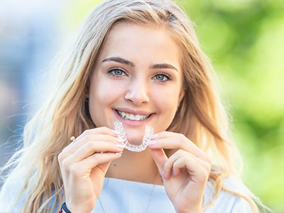 The image features a young woman with blonde hair and blue eyes, smiling and holding up a clear orthodontic aligner.