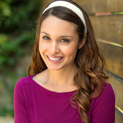 The image shows a woman with long brown hair, smiling at the camera. She is wearing a purple top and has a headband in her hair. The background consists of a wooden fence and greenery.