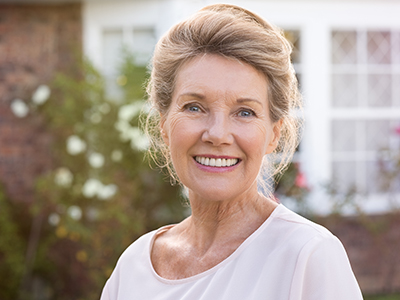 The image is a portrait of an older woman with short blonde hair, wearing a light pink top and standing outdoors in front of a house.