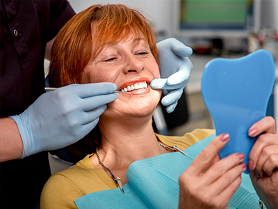 A woman in a dental chair receiving a teeth cleaning, with a blue toothbrush held up for inspection by a dental hygienist.