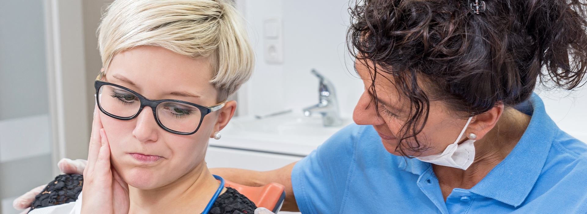 A woman with short hair is seated in a dental chair, receiving attention from a dentist who stands beside her.