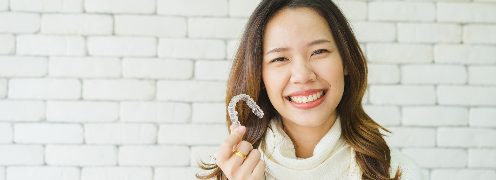 A woman with a radiant smile holds a ring, set against a white brick wall.