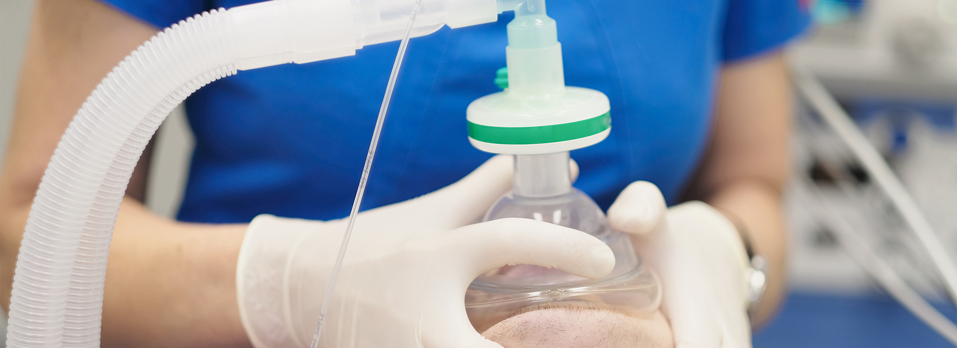 The image shows a person wearing gloves and a blue shirt, holding a clear plastic tube with a green cap attached to a transparent device, possibly a medical machine or equipment.