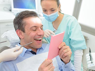 The image depicts a man in a dental office, holding up a pink card with a smile, surrounded by dental equipment and a female dental professional.