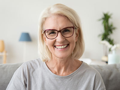 The image features a woman with short blonde hair, wearing glasses and a light-colored top, smiling at the camera. She appears to be sitting indoors with a neutral background that includes a lamp and some decorative items.