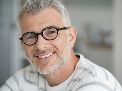 The image shows a man with gray hair and glasses, wearing a white shirt with a patterned collar. He has a beard and mustache, and is smiling at the camera.