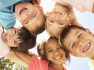 A group of children, both boys and girls, smiling and posing for a photo with their arms around each other, set against a bright outdoor background.