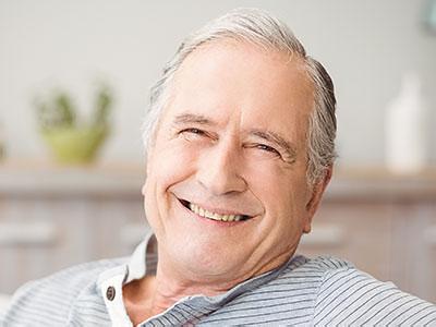 The image shows a man with gray hair, smiling and looking directly at the camera. He is wearing glasses and has a white beard. He appears to be sitting in a relaxed position, possibly in his home or a casual setting.