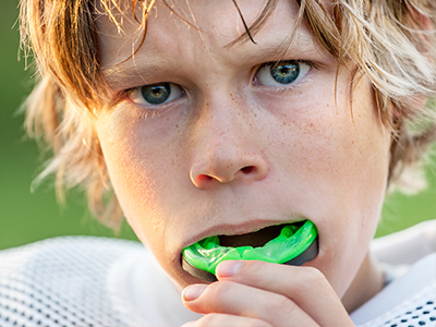 The image shows a young person with blonde hair and blue eyes, who appears to be a football player, holding a green object in their mouth while looking directly at the camera.