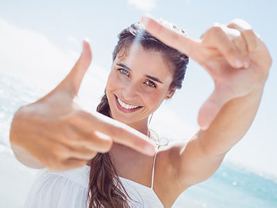 The image shows a woman taking a selfie with her hand and fingers forming the shape of a frame, set against a bright background that suggests a beach setting.